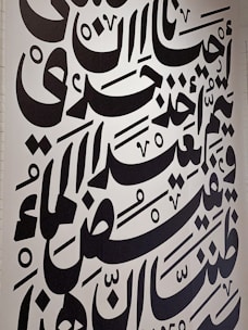 Large black Arabic script covers a white wall, featuring bold calligraphic letters with a variety of shapes and diacritical marks. The text is arranged in a visually dynamic and artistic manner, creating an intricate pattern.