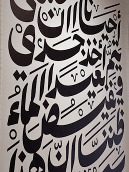 Large black Arabic script covers a white wall, featuring bold calligraphic letters with a variety of shapes and diacritical marks. The text is arranged in a visually dynamic and artistic manner, creating an intricate pattern.