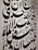 Large black Arabic script covers a white wall, featuring bold calligraphic letters with a variety of shapes and diacritical marks. The text is arranged in a visually dynamic and artistic manner, creating an intricate pattern.