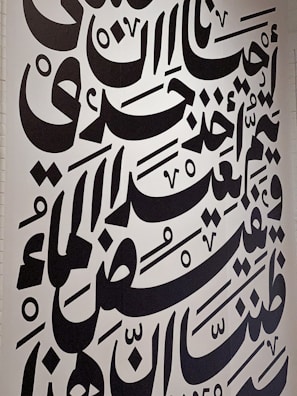 Large black Arabic script covers a white wall, featuring bold calligraphic letters with a variety of shapes and diacritical marks. The text is arranged in a visually dynamic and artistic manner, creating an intricate pattern.