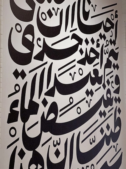 Large black Arabic script covers a white wall, featuring bold calligraphic letters with a variety of shapes and diacritical marks. The text is arranged in a visually dynamic and artistic manner, creating an intricate pattern.