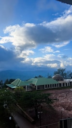 A landscape featuring a large building with green roofs, set against a backdrop of mountains and a partly cloudy sky. The foreground shows an open area with a few trees and individuals walking. The sky has a mix of white and dark clouds, suggesting a transitional weather pattern.