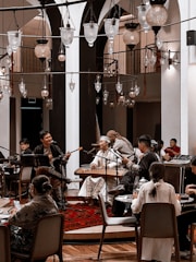 A cozy indoor setting with a group of musicians playing traditional instruments. The performers are dressed in cultural attire and are surrounded by an intimate audience seated at tables. Several ornate hanging lamps add a warm ambiance to the room, which features arches and wooden accents.