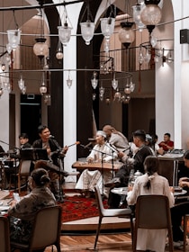 A cozy indoor setting with a group of musicians playing traditional instruments. The performers are dressed in cultural attire and are surrounded by an intimate audience seated at tables. Several ornate hanging lamps add a warm ambiance to the room, which features arches and wooden accents.