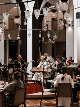 A cozy indoor setting with a group of musicians playing traditional instruments. The performers are dressed in cultural attire and are surrounded by an intimate audience seated at tables. Several ornate hanging lamps add a warm ambiance to the room, which features arches and wooden accents.