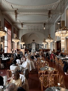 The image depicts a grand and ornate cafe or restaurant interior, featuring high ceilings with intricate moldings, large chandeliers, and wooden chairs and tables. Patrons are sitting and enjoying their meals, creating a lively atmosphere. The decor has a classic, luxurious feel with marble accents and rich wood tones.