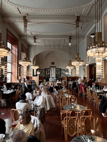 The image depicts a grand and ornate cafe or restaurant interior, featuring high ceilings with intricate moldings, large chandeliers, and wooden chairs and tables. Patrons are sitting and enjoying their meals, creating a lively atmosphere. The decor has a classic, luxurious feel with marble accents and rich wood tones.