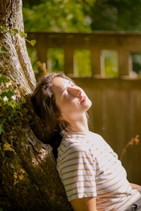 A person wearing a striped shirt leans back against a tree trunk, with closed eyes, seemingly enjoying the sunlight filtering through the foliage. The background features a wooden fence and greenery, giving a sense of relaxation and tranquility.