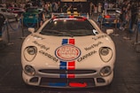 A white sports car with prominent logos including Hard Rock and Carrera is displayed indoors at an auto show. The car is surrounded by a blue rope barrier and is in front of a crowd of people viewing other vehicles.