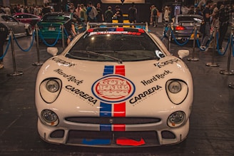 A white sports car with prominent logos including Hard Rock and Carrera is displayed indoors at an auto show. The car is surrounded by a blue rope barrier and is in front of a crowd of people viewing other vehicles.