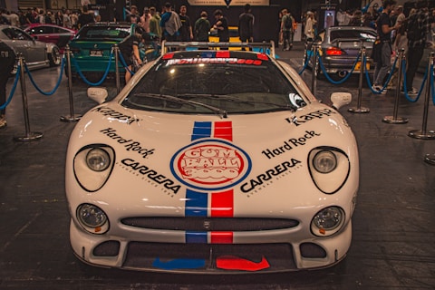 A white sports car with prominent logos including Hard Rock and Carrera is displayed indoors at an auto show. The car is surrounded by a blue rope barrier and is in front of a crowd of people viewing other vehicles.