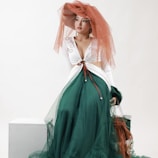 A woman seated wearing a fashion-forward outfit featuring a green voluminous skirt with sheer layers and a fitted white top adorned with delicate embroidery. The ensemble is accessorized with a large, dramatic orange hat that has translucent trailing fabric.