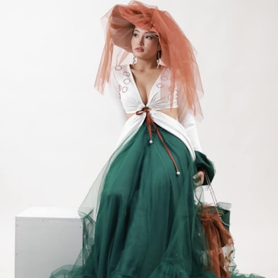 A woman seated wearing a fashion-forward outfit featuring a green voluminous skirt with sheer layers and a fitted white top adorned with delicate embroidery. The ensemble is accessorized with a large, dramatic orange hat that has translucent trailing fabric.