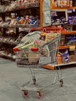 A shopping cart filled with a variety of packaged goods, including snacks and household items, is situated in an aisle of a grocery store. The shelves in the background are stocked with various colorful products.