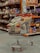 A shopping cart filled with a variety of packaged goods, including snacks and household items, is situated in an aisle of a grocery store. The shelves in the background are stocked with various colorful products.