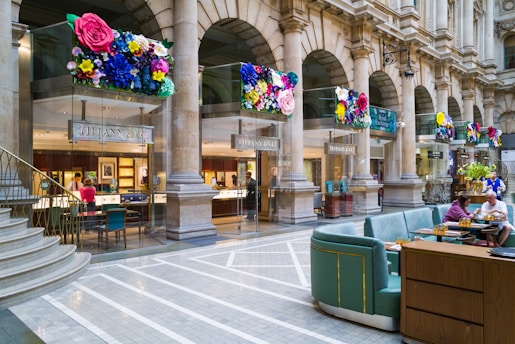 A luxurious shopping area featuring a Tiffany & Co store with elegant columns and decorative floral displays above the entrance. The interior of the jewelry store is visible, showcasing elegant displays and customers being assisted by staff. The adjacent area has comfortable seating with people enjoying refreshments. The setting is sophisticated and vibrant, combining the historical architecture with modern luxury.