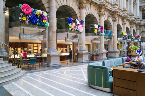 A luxurious shopping area featuring a Tiffany & Co store with elegant columns and decorative floral displays above the entrance. The interior of the jewelry store is visible, showcasing elegant displays and customers being assisted by staff. The adjacent area has comfortable seating with people enjoying refreshments. The setting is sophisticated and vibrant, combining the historical architecture with modern luxury.