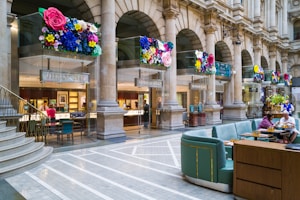 A luxurious shopping area featuring a Tiffany & Co store with elegant columns and decorative floral displays above the entrance. The interior of the jewelry store is visible, showcasing elegant displays and customers being assisted by staff. The adjacent area has comfortable seating with people enjoying refreshments. The setting is sophisticated and vibrant, combining the historical architecture with modern luxury.
