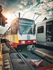 A tram with a red and yellow exterior approached a busy station marked by a sign indicating 'U74 Düsseldorf Hbf.' The sky is partly cloudy with the sun visible, casting an orange glow. Adjacent to the tram is a modern white train with a stroller design symbol on the side. The area is lined with rails and pedestrian walkway markings.