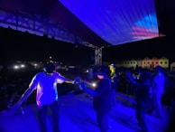 A live music performance on stage during nighttime with several band members playing instruments. The stage is illuminated with blue and purple lights, and there is a visible audience in the background.