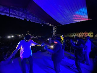 A live music performance on stage during nighttime with several band members playing instruments. The stage is illuminated with blue and purple lights, and there is a visible audience in the background.
