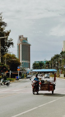A street scene featuring a person riding a motorized cart or scooter with a canopy, likely used for selling food or goods. In the background, there is a large, modern building with a distinctive architectural design. Several trees and utility poles line the street, and other vehicles, including motorcycles, are also present.