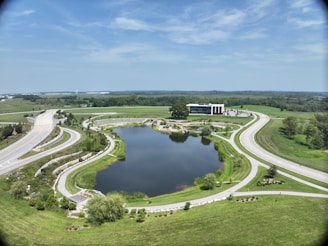 A wide landscape featuring a large man-made pond surrounded by well-maintained greenery and roads forming circular patterns. A modern building is situated near the pond, with open fields and trees in the background. The sky is clear with a few scattered clouds.