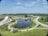 A wide landscape featuring a large man-made pond surrounded by well-maintained greenery and roads forming circular patterns. A modern building is situated near the pond, with open fields and trees in the background. The sky is clear with a few scattered clouds.