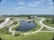 A wide landscape featuring a large man-made pond surrounded by well-maintained greenery and roads forming circular patterns. A modern building is situated near the pond, with open fields and trees in the background. The sky is clear with a few scattered clouds.