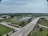 An aerial view of a landscape featuring a large building on the left, a park area with a pond at the center, and roads curving around the area. The setting is lush with green fields, and there are trees scattered throughout the scene. The sky is partly cloudy, adding a serene backdrop to the image.