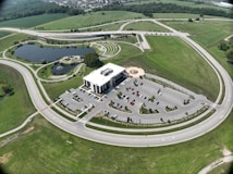 An aerial view of a large, modern building surrounded by an expansive parking lot with numerous cars parked. The area is circled by a wide road, and there is lush green grass covering the surrounding landscape. A small pond with a fountain and landscaped areas featuring pathways and greenery are visible. The setting conveys a sense of organization and spaciousness.