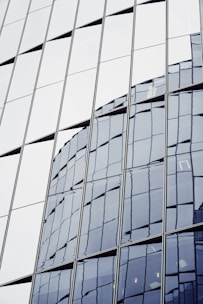 Reflective glass windows of a modern building, showing a distorted reflection of another skyscraper. The angular panels create a geometric pattern.