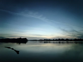 A slow-moving river reflecting muted tones of twilight, surrounded by sparse trees.