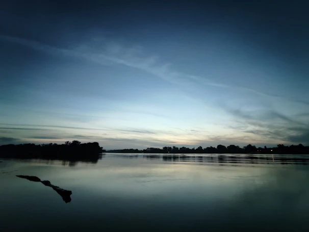 A slow-moving river reflecting muted tones of twilight, surrounded by sparse trees.