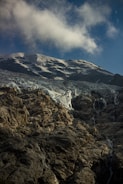 A rugged mountain landscape with snow-covered peaks under a clear blue sky. Wispy clouds drift above the rocky, textured surface of the mountains. The glacier exhibits layers of white ice mingling with darker rock formations.