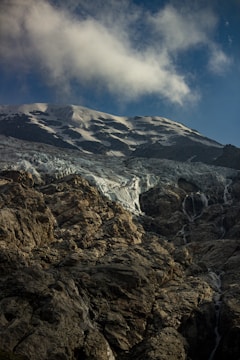 A rugged mountain landscape with snow-covered peaks under a clear blue sky. Wispy clouds drift above the rocky, textured surface of the mountains. The glacier exhibits layers of white ice mingling with darker rock formations.