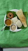 A plate of South Indian breakfast items is presented, featuring a dosa, idlis, a vada, sambar in a small bowl, and coconut chutney in another bowl, all served on a banana leaf over a white plate. The setting is against a green fabric background, showcasing traditional Indian cuisine.