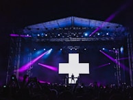 A concert stage is set outdoors at night with bright purple and blue lights illuminating the scene. A large LED screen displays a white plus sign, and a silhouette of a performer stands in front of it. The crowd is visible in the foreground, with several people holding up phones to capture the performance.