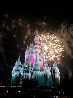 A large, illuminated castle adorned with colorful lights is set against a dark night sky. Vibrant fireworks burst overhead, creating a festive and magical atmosphere. The structure features multiple spires and is highlighted by bright blue and pink hues.