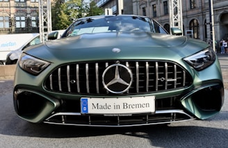 A front view of a stylish green Mercedes-Benz car with a prominent grille and sleek design. The car features a license plate that reads 'Made in Bremen.' The background includes a building with arched windows and a partially visible white van.