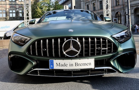 A front view of a stylish green Mercedes-Benz car with a prominent grille and sleek design. The car features a license plate that reads 'Made in Bremen.' The background includes a building with arched windows and a partially visible white van.