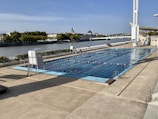 An outdoor swimming pool is situated next to a large river, with several people swimming in designated lanes. Surrounding the pool is a spacious concrete deck area with a lifeguard chair. Trees and buildings can be seen across the river, along with a bridge in the background. The sky is clear with a few clouds.