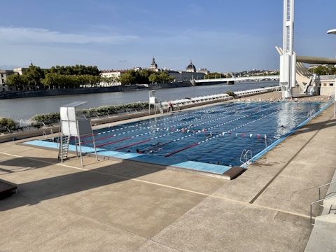 An outdoor swimming pool is situated next to a large river, with several people swimming in designated lanes. Surrounding the pool is a spacious concrete deck area with a lifeguard chair. Trees and buildings can be seen across the river, along with a bridge in the background. The sky is clear with a few clouds.