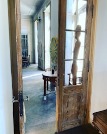 An open wooden door leads into a room featuring wooden furniture and artistic decorations. A wooden sculpture stands near the entrance, and there are vibrant circular art pieces on the walls. Chairs and tables are situated in a well-lit corridor, adding to the artistic and welcoming atmosphere.