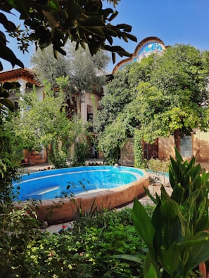 A lush garden surrounds a small, oval-shaped pool, with dense greenery including various plants and trees. The pool is empty and painted bright blue. The setting is enclosed within a courtyard featuring traditional architecture with wooden beams and a colorful decorative element visible above a building.
