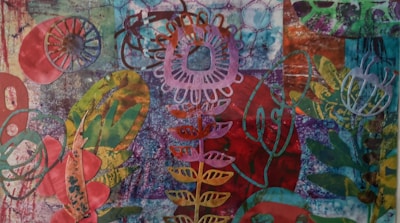 An abstract, colorful collage featuring various botanical patterns and shapes. The image includes floral motifs with intricate cut-outs placed on a textured background combining shades of blue, red, green, and purple.