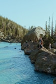 A serene lakeside scene surrounded by lush evergreen trees and rocky terrain. Several people are gathered along the edge of the water, some standing on large rocks, indicating a leisurely outdoor activity.