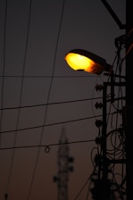 Silhouette of a street lamp pole with illuminating light against a dim sky background. Several power cables and electrical components are visible, creating an intricate pattern against the sky. In the distance, there is a blurred structure resembling a communication tower.