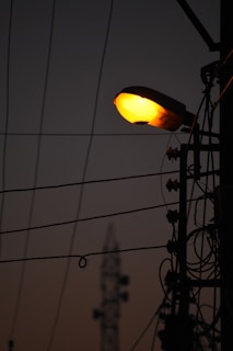 Silhouette of a street lamp pole with illuminating light against a dim sky background. Several power cables and electrical components are visible, creating an intricate pattern against the sky. In the distance, there is a blurred structure resembling a communication tower.