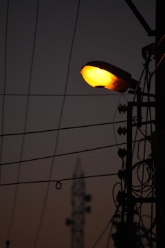 Silhouette of a street lamp pole with illuminating light against a dim sky background. Several power cables and electrical components are visible, creating an intricate pattern against the sky. In the distance, there is a blurred structure resembling a communication tower.